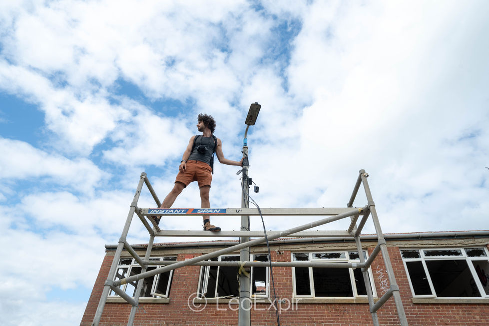 Person stands on scaffolding in front of an abandoned brick building, holding onto a lamp post against the bright blue, cloud-filled sky. Fraser Range, Portsmouth.
