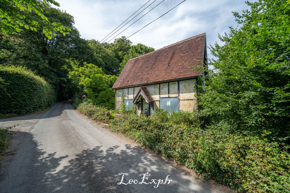 A rustic abandoned cottage with a steep red-tiled roof sits amidst lush greenery by a narrow country road, evoking a tranquil, idyllic countryside scene.