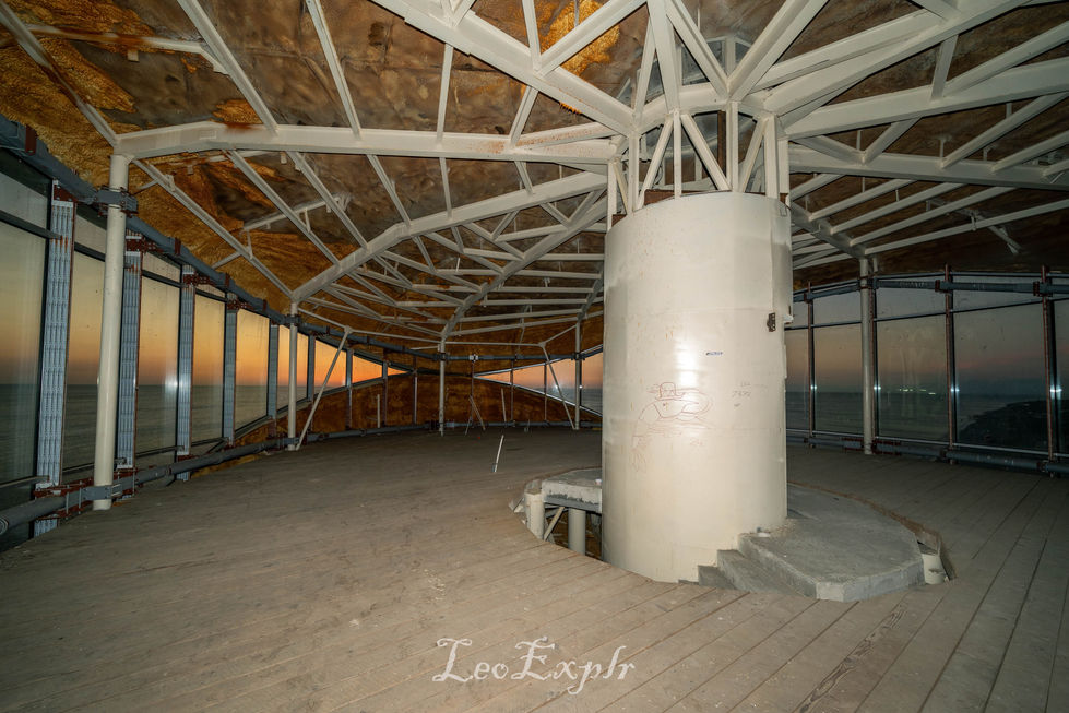 Interior of an unfinished circular building with a white central column, metallic framework, and large windows overlooking a sunset. Sparse and industrial. Anaklia abandoned tower in Georgia.