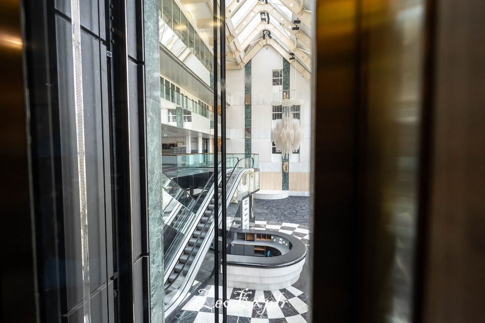 View from a glass elevator overlooking a modern, spacious atrium. Features an escalator, geometric chandelier, and checkered floor, creating an elegant, airy ambiance.