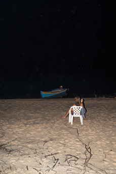 A person sits in a white chair on a sandy beach at night, gazing at a blue boat on the dark water. The scene is calm and introspective.