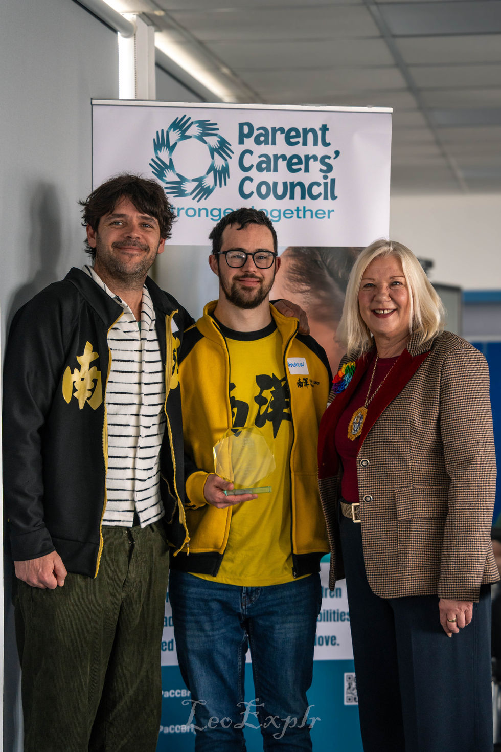 Three people stand smiling in front of a "Parent Carers' Council" banner. The person in the center holds an award, and they appear joyful and proud. On the right stands a woman, the mayor of Brighton & Hove.