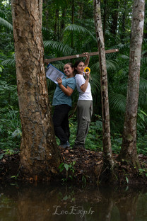 Two women in a lush forest stand back-to-back between trees, one holding papers and smiling, the other with a measuring tape, displaying camaraderie.