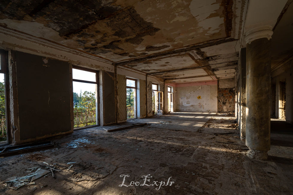 Inside the abandoned Sanatorium Shaktar, also known as Sanatorium Meshakhte, in Tskaltubo in Georgia. Abandoned, sunlit room with peeling walls and exposed concrete columns. Large windows reveal overgrown greenery outside, casting long shadows inside. Atmosphere is desolate.