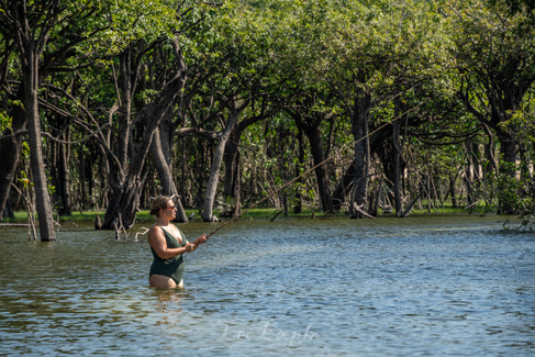 A person standing waist-deep in water, fishing with a rod in a sunlit, dense mangrove forest. The scene is serene and natural. Amazon Rainforest, Tapajos River.