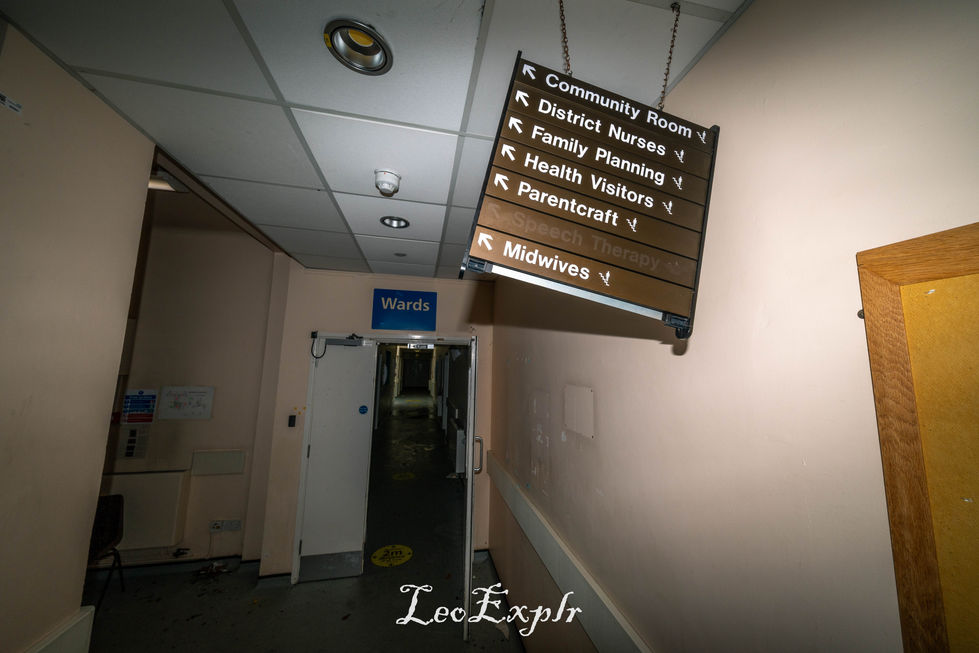 Dimly lit hospital corridor with a directory sign listing rooms like "Community Room" and "Midwives." An open door labeled "Wards" is in the background. Abandoned Edenbridge and District War Memorial Hospital.