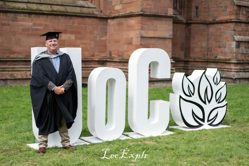 A graduate in cap and gown stands smiling beside large white letters "UoC" for University of Carlisle and a leaf emblem. The backdrop features the rustic red brick wall of Carlisle Cathedral, conveying achievement and celebration.