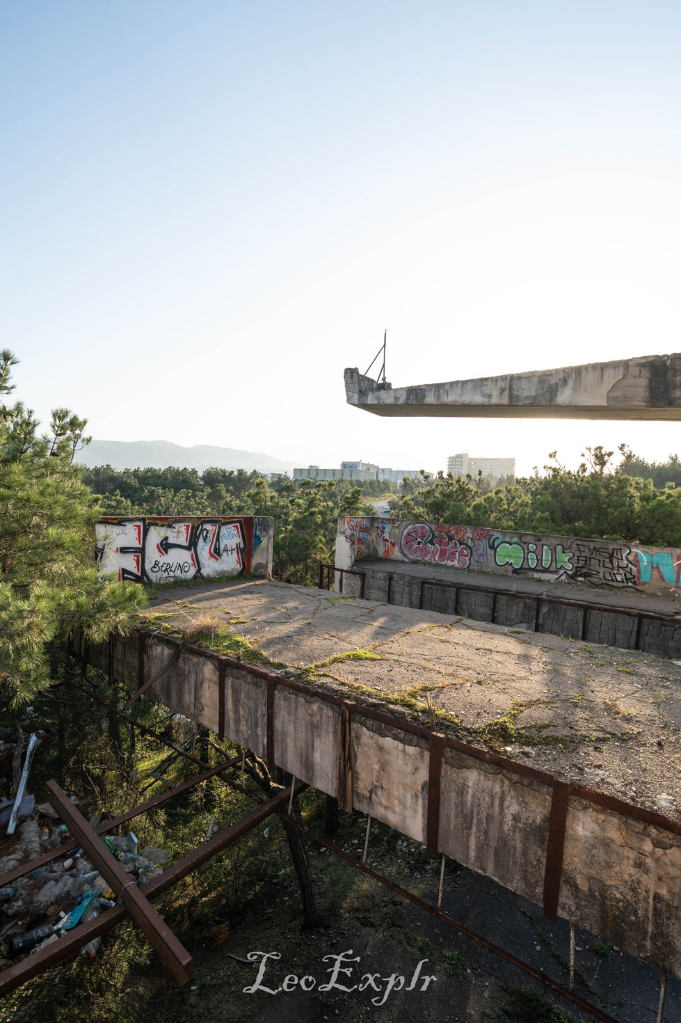 Abandoned concrete structure with rusty beams and graffiti. Overgrown with weeds, it overlooks distant buildings and trees under a clear sky. Former Lisi Tramway Cable Car Station in Tbilisi.