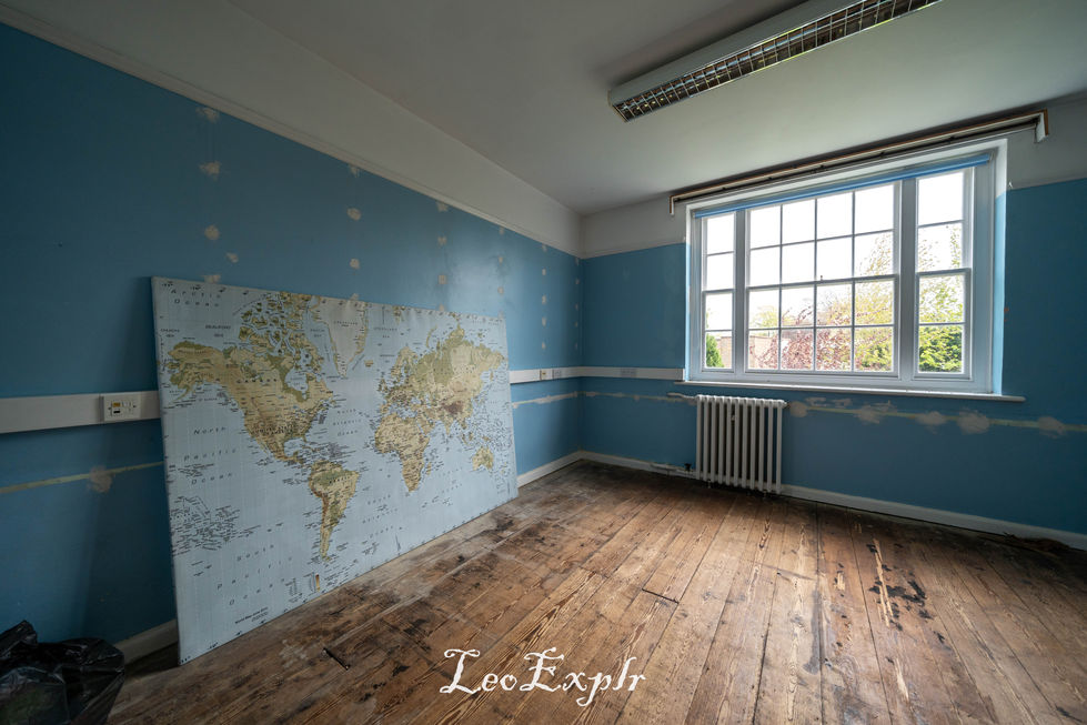 An empty room with pale blue walls and a world map leaning against one wall. The floor is wooden and worn. A large window offers natural light. Inside Steyning Grammar School abandoned Church Street site, Sussex.