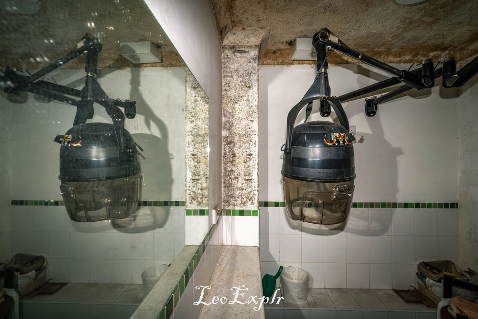 A vintage wall-mounted hair dryer with a cracked ceiling is reflected in a dirty mirror. Surroundings include tiled walls and scattered cleaning items, suggesting neglect. Inside an abandoned care home. Clemsfold House, Horsham.