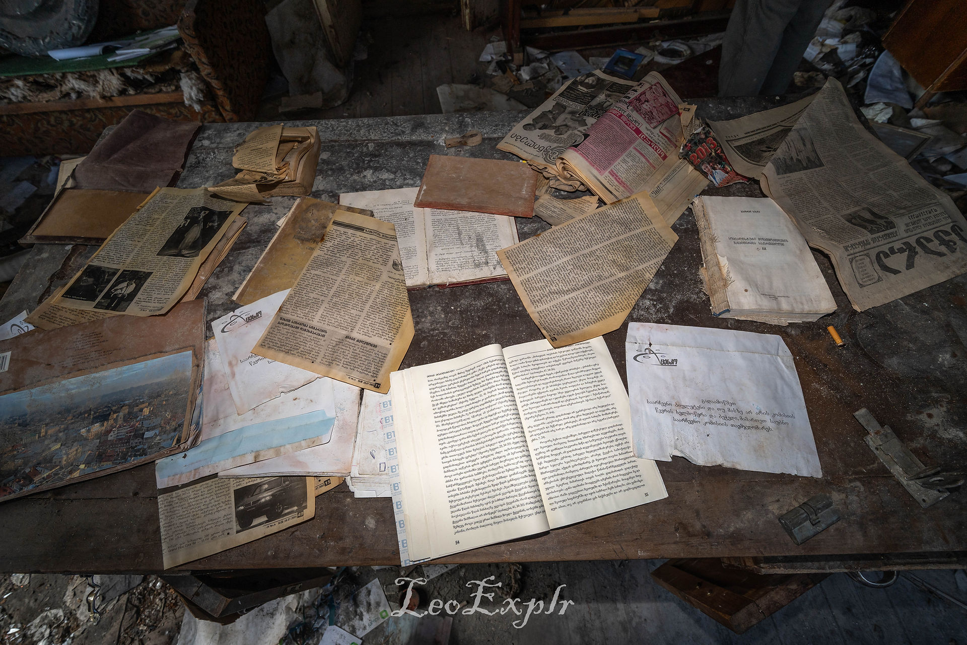 Scattered old newspapers, documents, and an open book cover a dusty tabletop. The scene conveys a sense of history and abandonment. Former Palace of Culture in Dagva, Georgia.