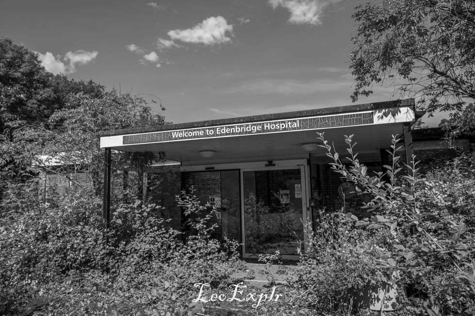 Black and white photo of an overgrown, abandoned building with a "Welcome to Edenbridge Hospital" sign. The scene feels desolate and neglected.