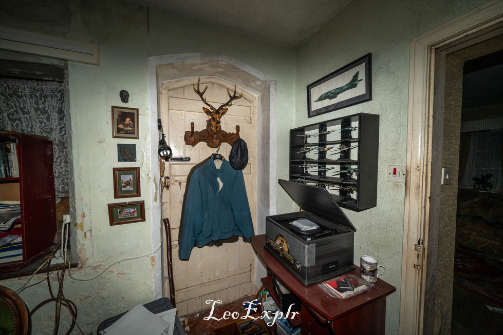 Dimly lit room with cluttered shelves, a coat hanging on a deer head hook, retro audio equipment on a table, and aviation art and models on the wall.