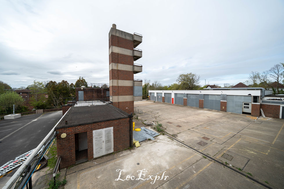 Abandoned brick building of a former fire station with narrow tower, surrounded by empty concrete lots. Overcast sky, trees in the background, conveying a desolate atmosphere.