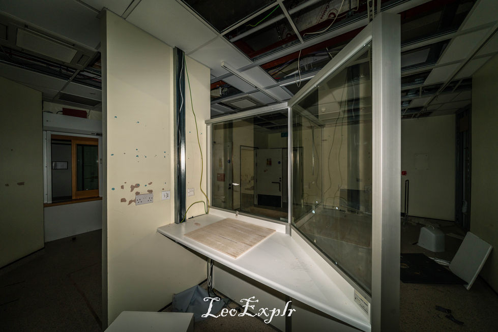Abandoned interior of an x-ray room with a broken ceiling and exposed wires inside an abandoned hospital. A dusty counter with glass panels is in the foreground, creating a neglected, eerie atmosphere. Edenbridge and District War Memorial Hospital.