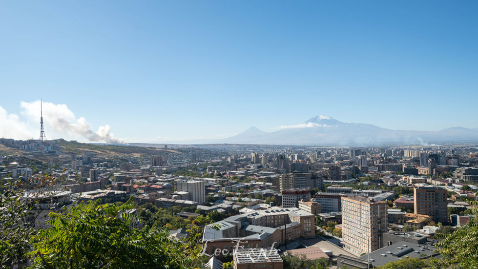 Panoramic view of Yerevan, Armenia, under a clear blue sky. A TV tower is on the left, with Mount Ararat in the background. Buildings and greenery fill the foreground.