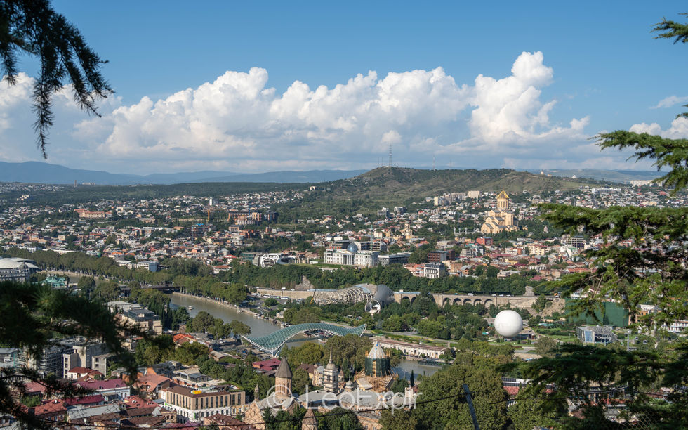 Panoramic cityscape of Tbilisi, Georgia, with the Kura River, Peace Bridge, hillside homes, church domes, and bright blue sky.
