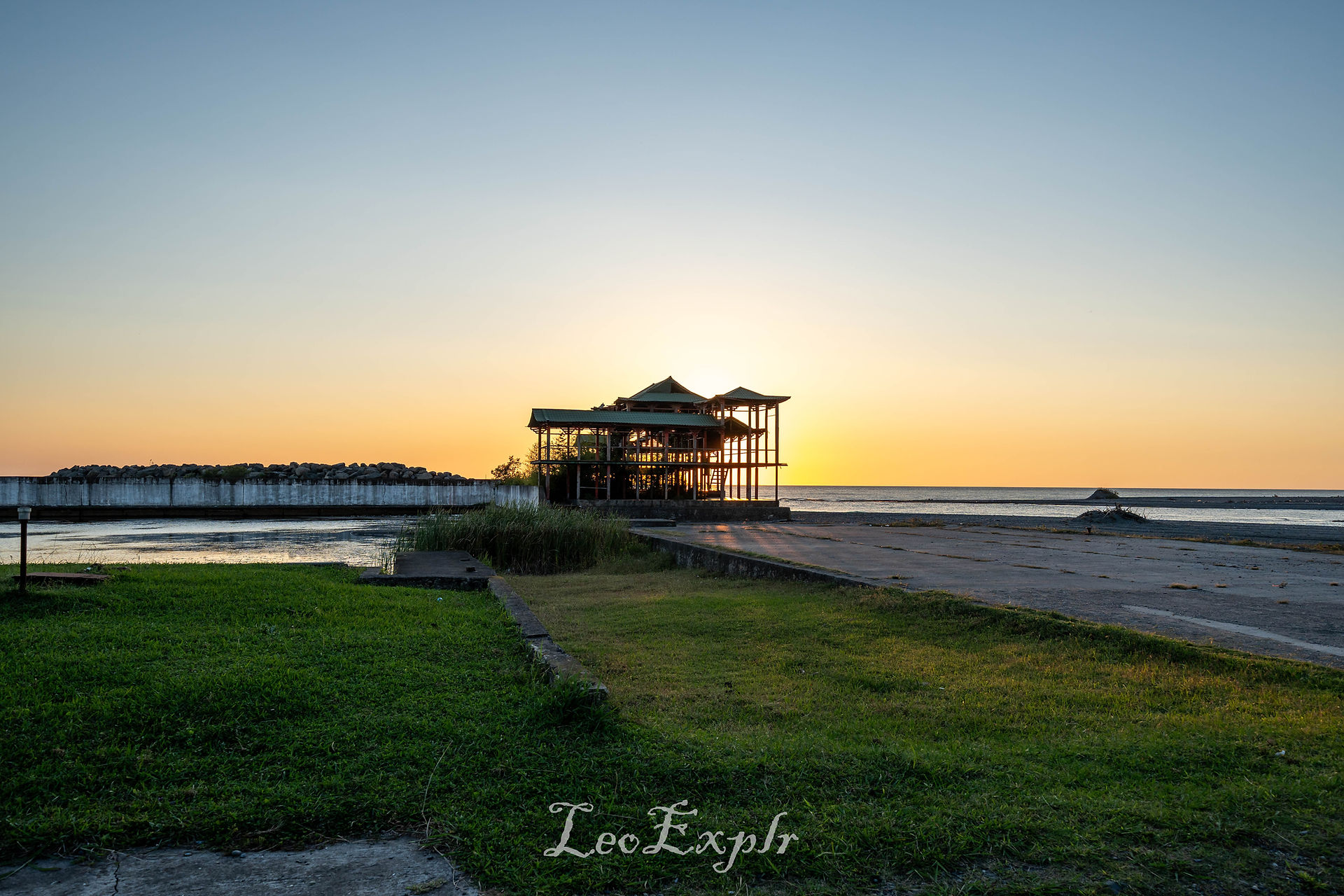 Anaklia Beach in Georgia. Silhouette of an unfinished beachside structure at sunset, surrounded by calm sea. The horizon glows with warm orange hues, contrasting with grassy foreground.