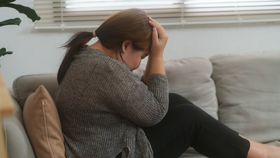 Woman sits on a couch with her head in her hands, looking stressed in a bright living room with blinds and pillows.