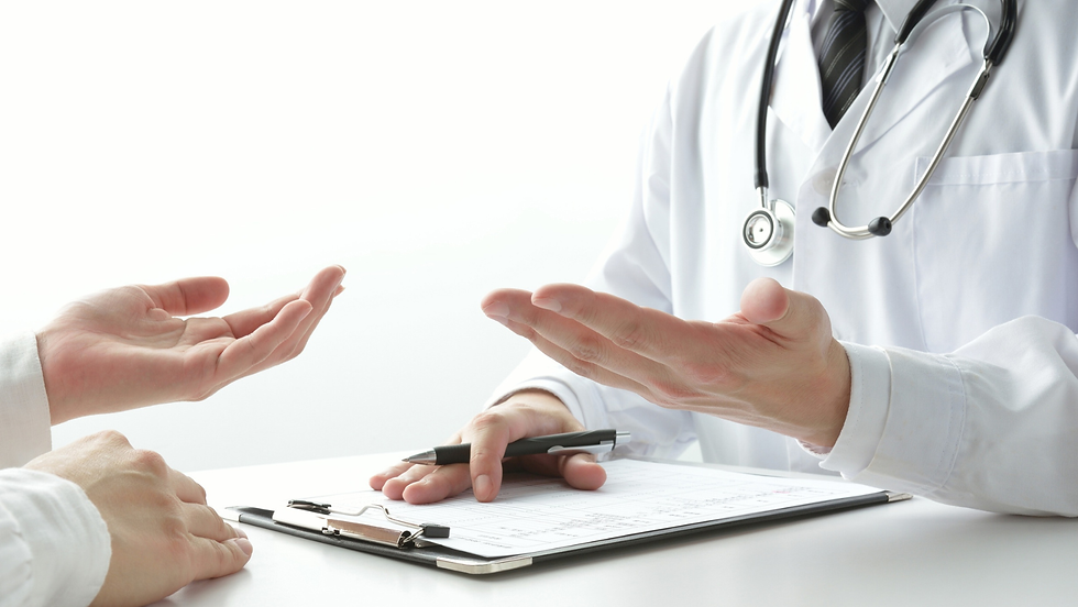 Doctor in white coat with stethoscope gestures during a consultation, facing a patient’s open hands over a clipboard in bright clinic.