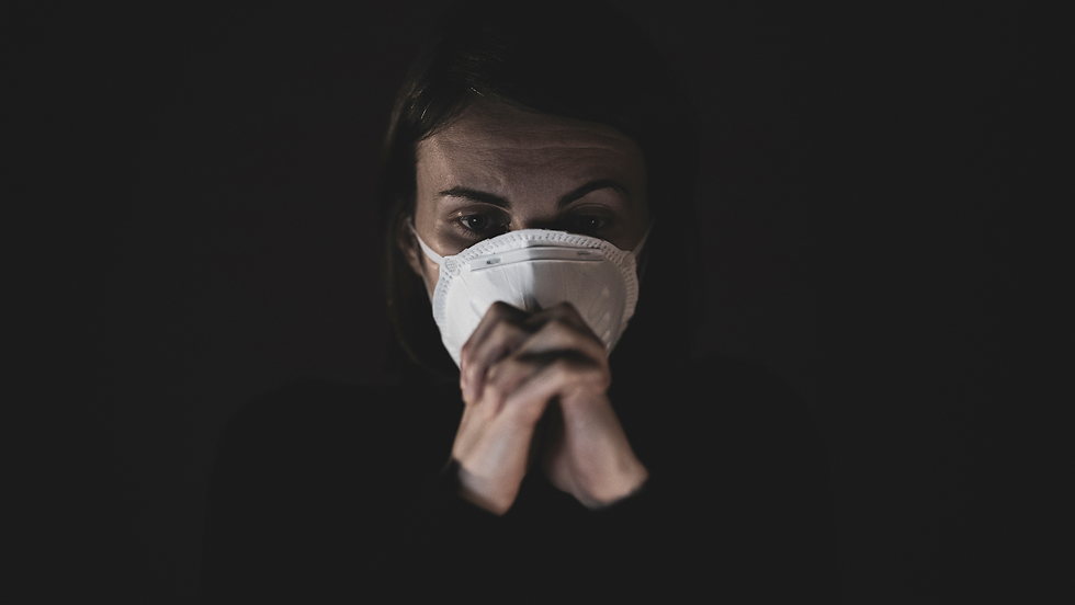 Masked woman with clasped hands stares ahead in dim light against a dark background, conveying fear or worry.