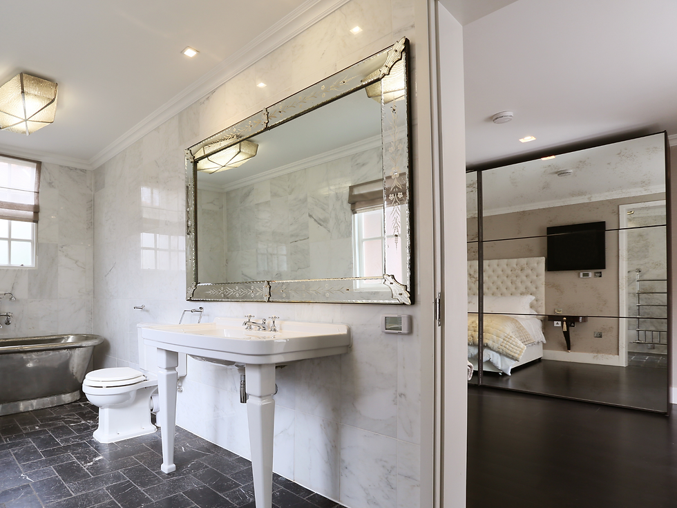 Luxurious marble bathroom with freestanding tub, pedestal sink and large ornate mirror; bedroom reflected in mirrored closet doors.