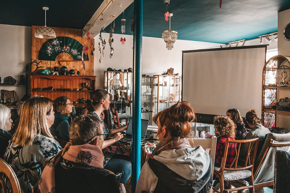 Many people young and old are seated on wooden chairs inside a crystal shop facing a large white screen. A blue pole sits in the centre of all those who are seated. Behind the pole is a lady on her laptop that is connected to a projector. They are waiting to watch a documentary film on the Crystal Shop and it's founder that they are sat in.