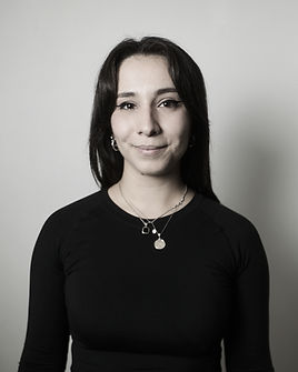 Portrait of Sol, a smiling tattoo artist with short hair, glasses, and a tropical-patterned shirt, photographed in black and