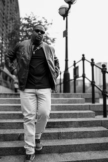 Black male model wearing a leather jacket and sunglasses walking down urban concrete steps in a confident street-style pose — fashion shoot in black and white.