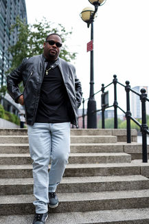 Black male model wearing a leather jacket and sunglasses walking down urban concrete steps in a confident street-style pose next to the canal — fashion shoot in colour.
