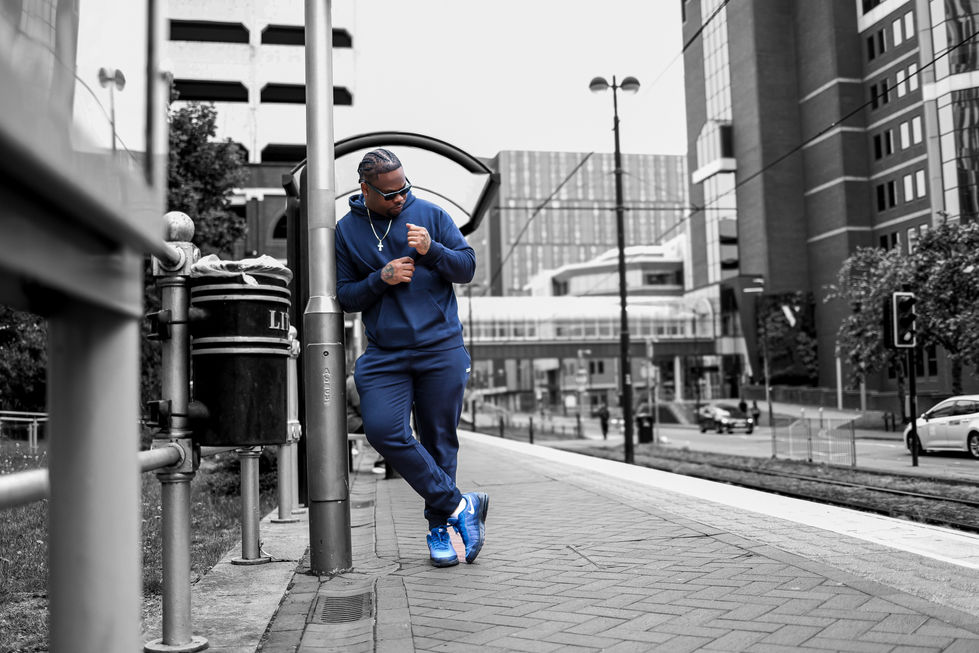 Black male model leaning casually against a post in a modern cityscape at the tram stop, dressed in a dark blue Nike tracksuit and sunglasses — urban fashion photoshoot in colour and black & white.