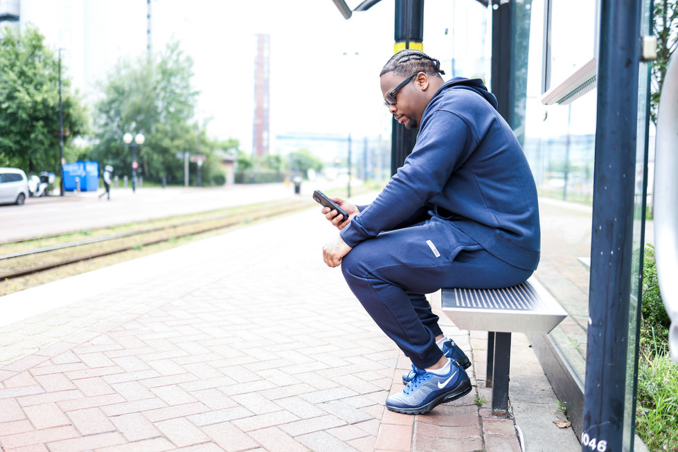 Black male model sitting casually at the tram stop looking at his phone, dressed in a dark blue Nike tracksuit and sunglasses — urban fashion photoshoot in colour.