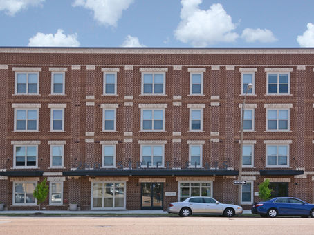 Red brick apartment building with white trim and large windows. Two parked cars in front, blue sky and clouds above. Street sign visible that reads "2ND STREET FLATS"