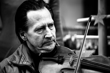 Black-and-white photo of a man playing a violin on the street with eyes closed, expression serene and emotional. The image evokes curiosity about his life and the beauty of his music.