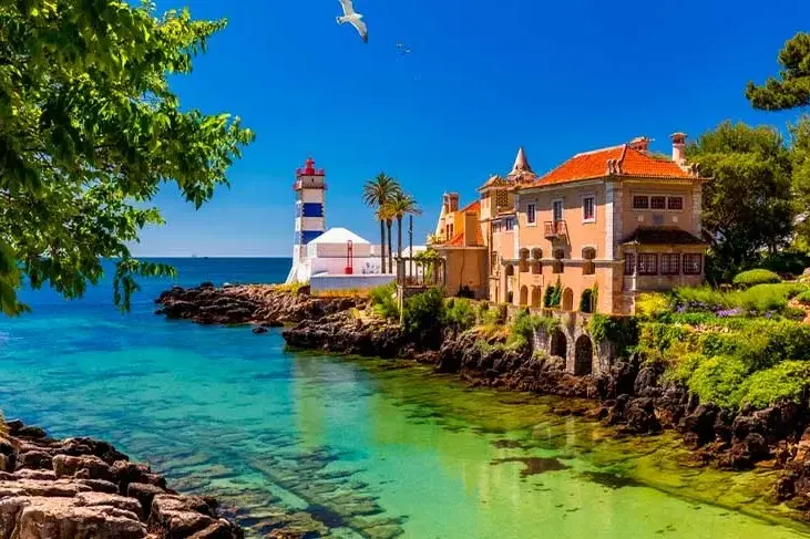 Colorful seaside villa and striped lighthouse overlook turquoise water under a bright blue sky, with trees and rocky shore of Caiscais
