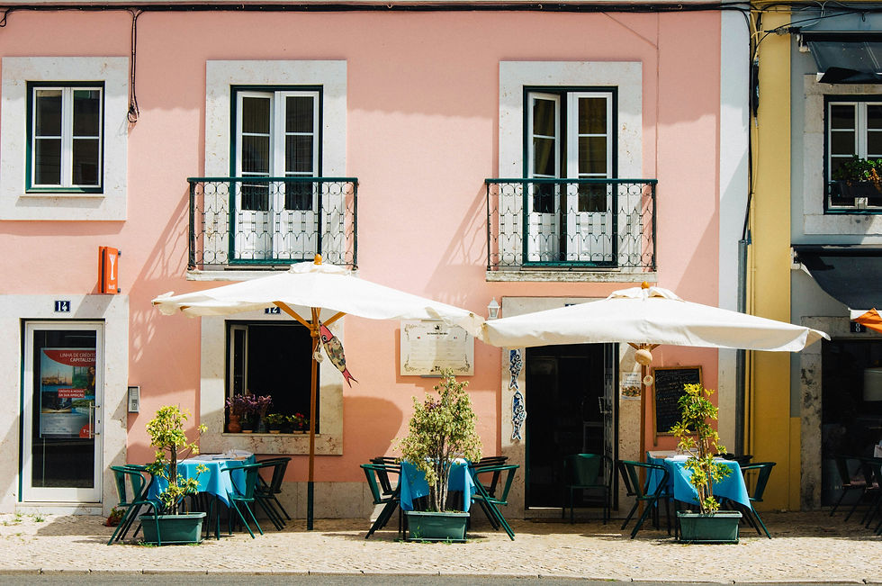 Pastel pink café facade with white umbrellas, blue tablecloths, potted plants, balconies, and a sunny quiet street scene in Lisbon