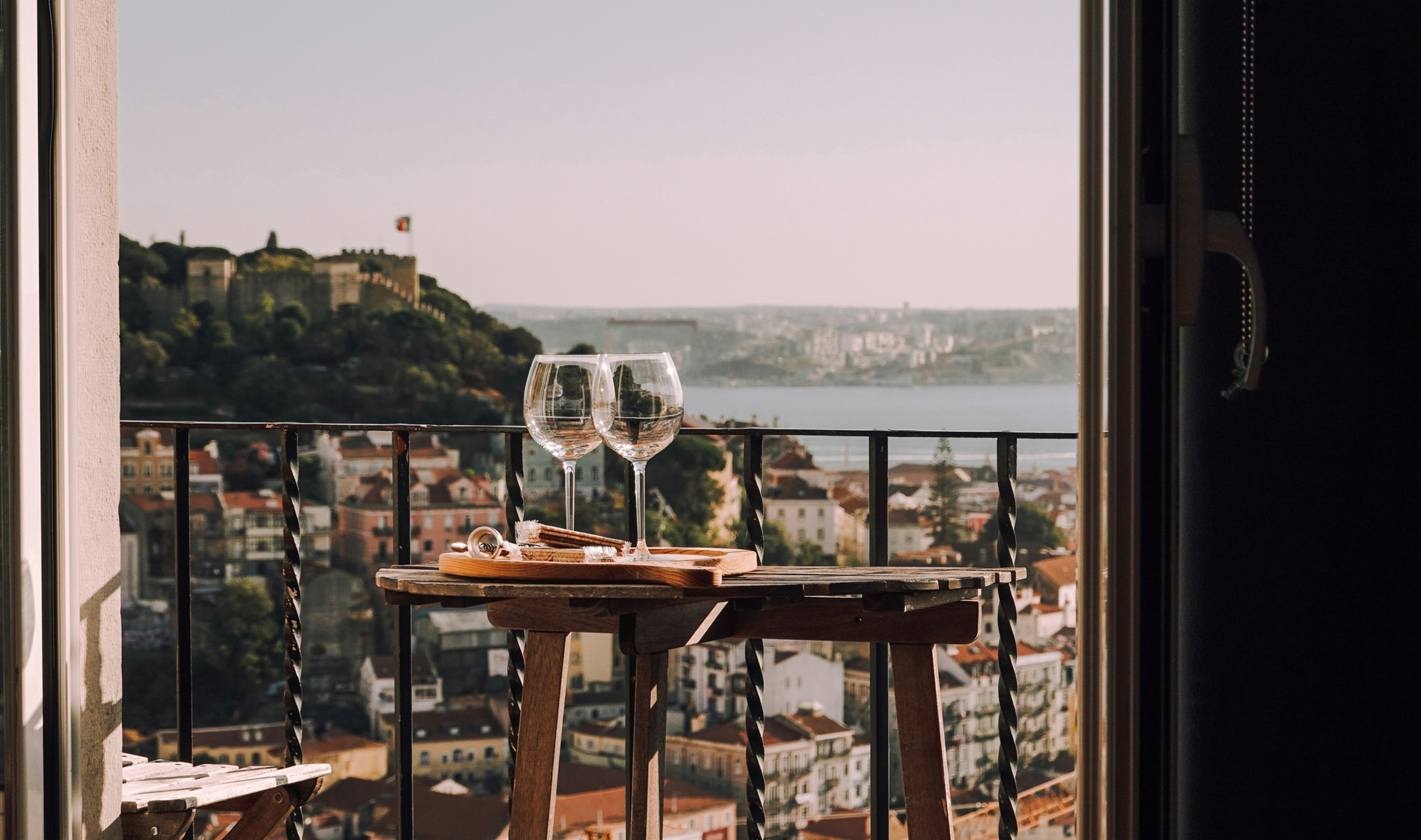 Two wine glasses on a balcony table overlook a sunlit city and river, creating a calm, romantic evening scene.