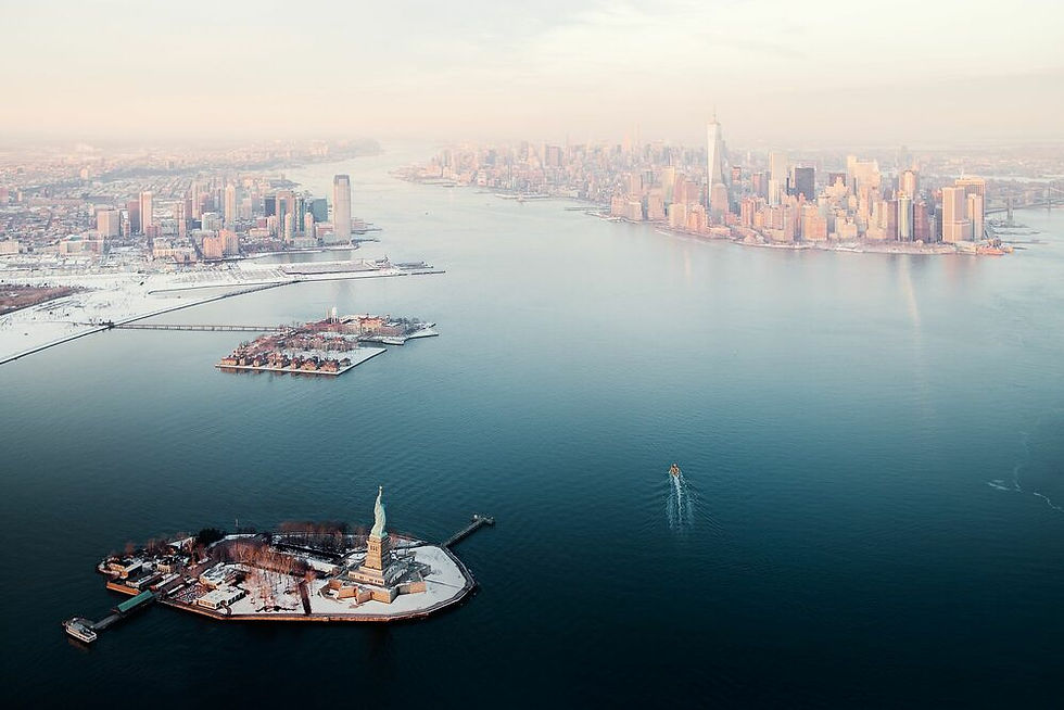 Aerial view of the Statue of Liberty and Manhattan skyline across New York Harbor, with a boat wake in calm water.