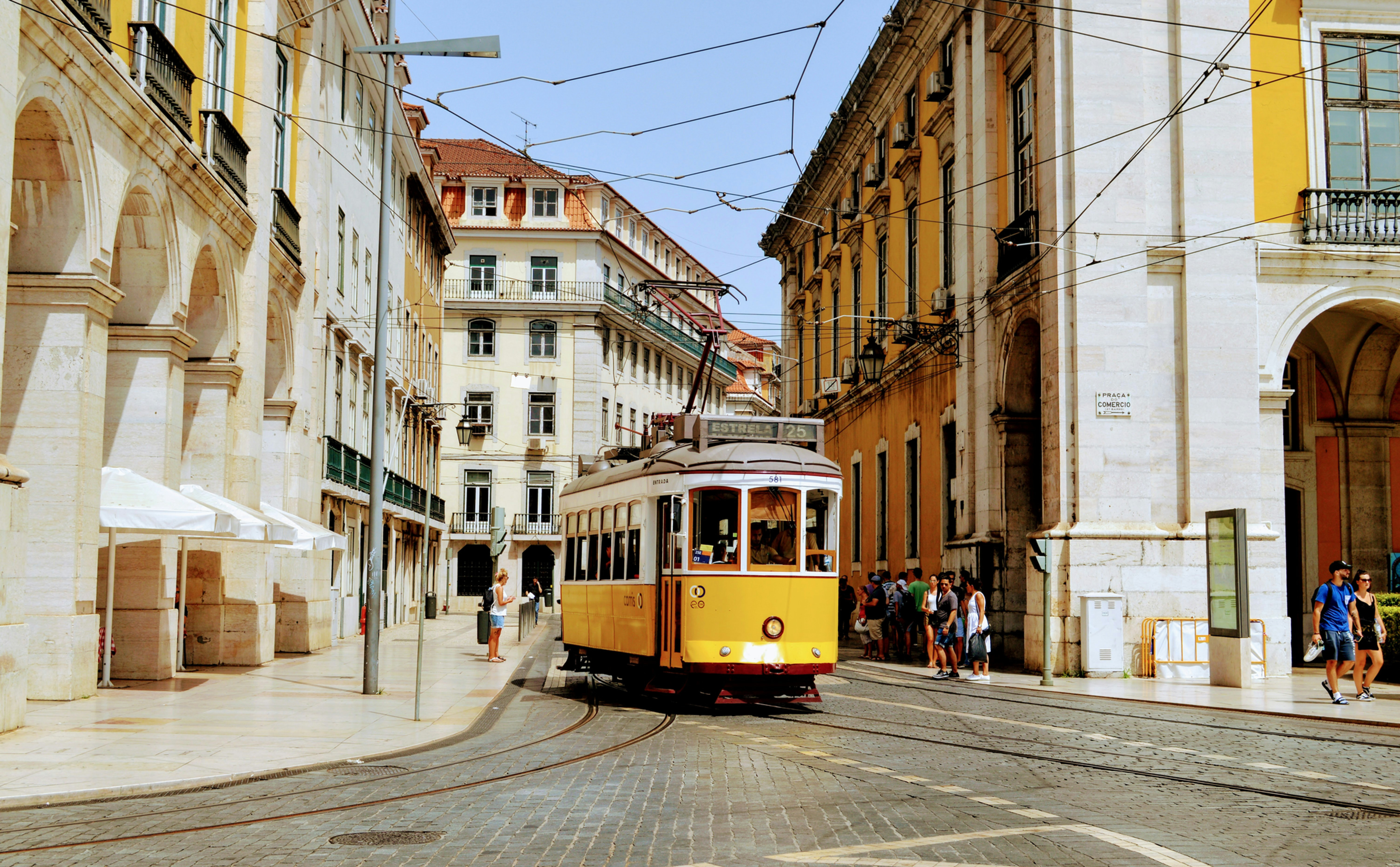 Yellow tram 25 passes through a sunny Lisbon street lined with historic buildings and pedestrians.