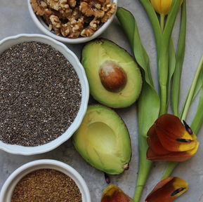 Flat lay of anti-inflammatory, hormone-supportive  foods- walnuts, chia sees, halved avocado and ground flaxseed in white bowls with yellow and orange tulips, from Balanced Imogen's food-first guide to anti-inflammatory foods for women's hormone health on The Balanced Edit. 