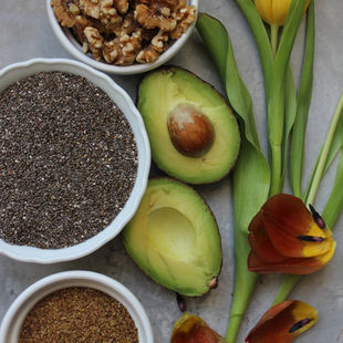 Flat lay of anti-inflammatory, hormone-supportive  foods- walnuts, chia sees, halved avocado and ground flaxseed in white bowls with yellow and orange tulips, from Balanced Imogen's food-first guide to anti-inflammatory foods for women's hormone health on The Balanced Edit. 