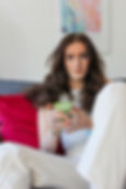 Balanced Imogen sitting on a sofa with colourful cushions, wearing a grey vest top and cream jeans, holding a matcha latte in a relaxed, natural setting.
