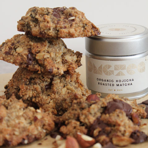 Balanced Imogen's hojicha nutty cookie clusters stacked on parchment paper alongside an organic hojicha roasted matcha tin- a hormone-supportive, cycle-synced luteal phase snack made with dark chocolate, ground almonds and pumpkin seeds.