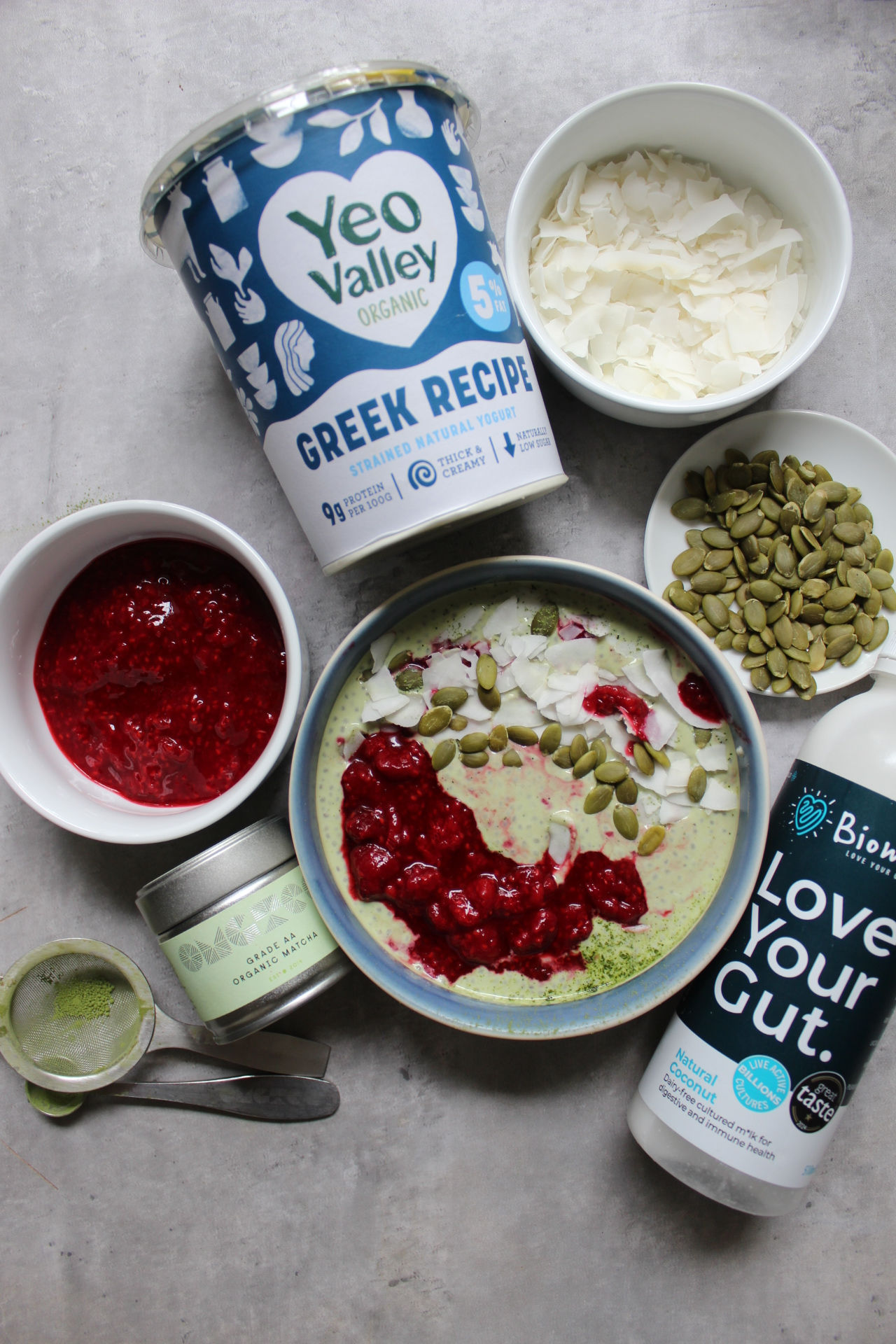 Overhead flat lay of Balanced Imogen's creamy matcha and raspberry chia pudding- styled with Yeo valley Greek yoghurt, Biomel coconut kefir, grade AA Organic OMG Matcha Tea, pumpkin seeds, coconut shavings and homemade raspberry compote, a cycle-synced breakfast for the follicular and ovulatory phases. 