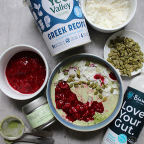 Overhead flat lay of Balanced Imogen's creamy matcha and raspberry chia pudding- styled with Yeo valley Greek yoghurt, Biomel coconut kefir, grade AA Organic OMG Matcha Tea, pumpkin seeds, coconut shavings and homemade raspberry compote, a cycle-synced breakfast for the follicular and ovulatory phases. 