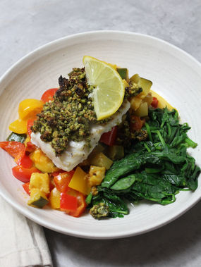 Pistachio parmesan crusted cod served with colourful ratatouille vegetables and fresh spinach and lemon on a white plate.