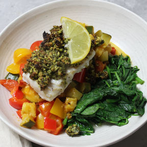 Pistachio parmesan crusted cod served with colourful ratatouille vegetables and fresh spinach and lemon on a white plate.