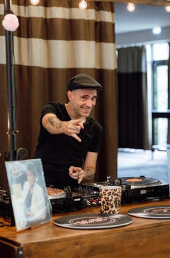 Un DJ souriant portant un béret noir et un tee-shirt noir, avec des tatouages sur les bras, pointe du doigt vers l'avant. Il se tient derrière une platine vinyle en bois dans une salle avec des rideaux à rayures marron et beige. Au premier plan, on voit une pochette d'album de Michael Jackson (Thriller) et un gobelet léopard.