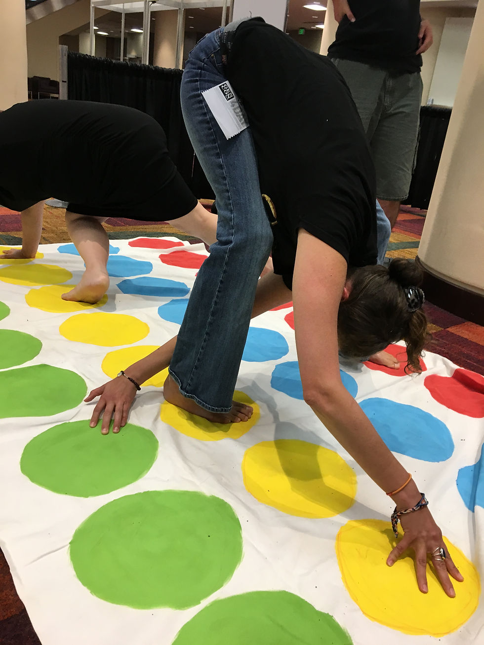 Joanna Stevens actor - playful Twister floor game action shot with right foot on yellow, left foot on red, left arm reaching through legs to a green dot and right arm stretched to a yellow dot, head bent low, bottom high in air, and colorful game mat clearly visible