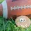 Thumbnail: close up of a football shaped dog cookie decorated with a smiling face sitting in front of a football on turf
